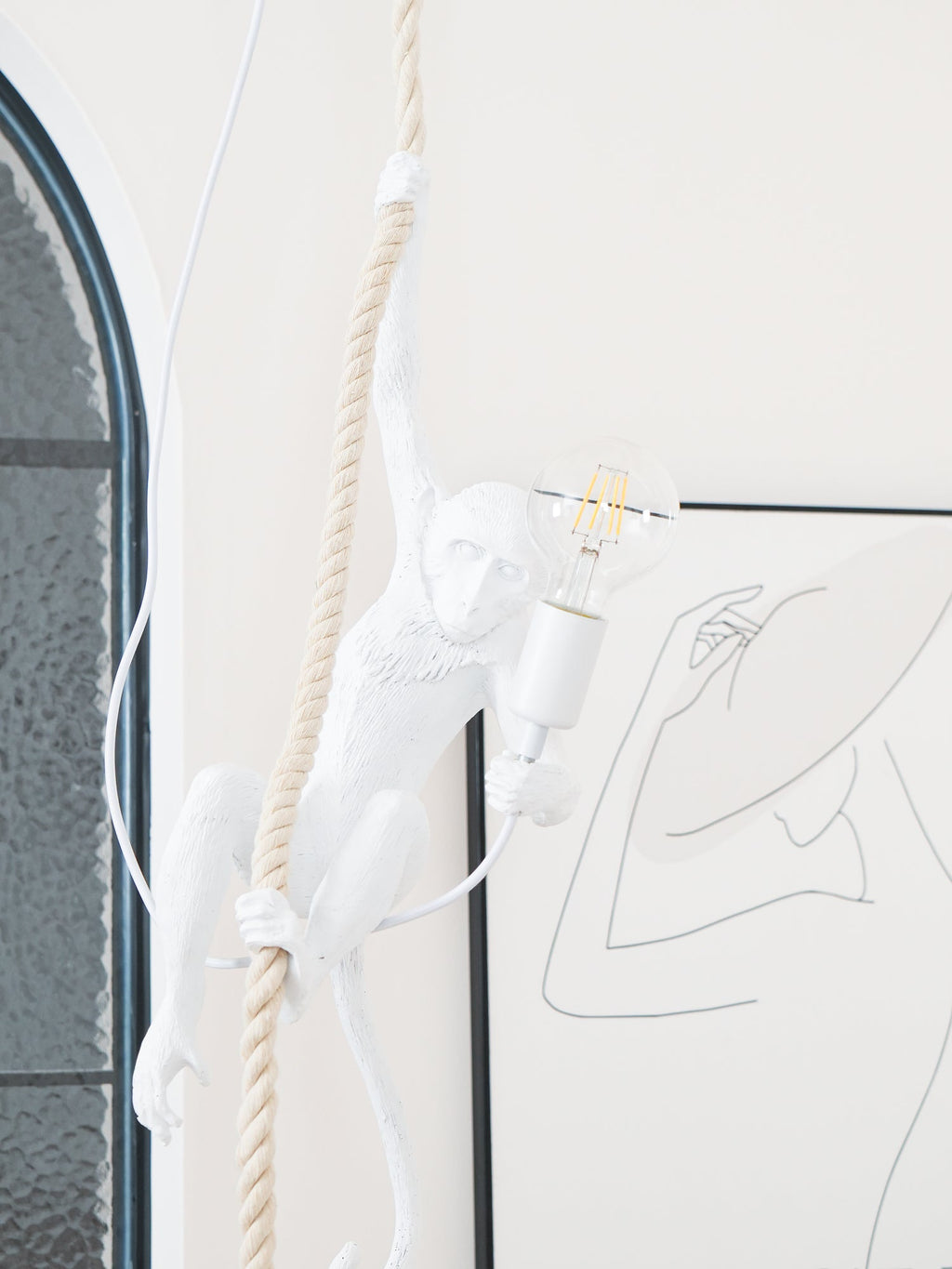 Narrow view of the white monkey gripping the rope with a filament bulb lit, in front of an arched window; from Clérachic.