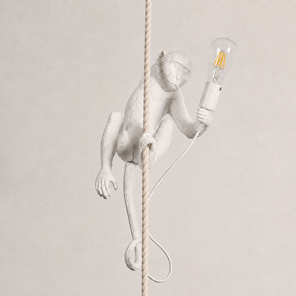 White monkey wrapped around a thick rope on a soft beige wall, holding a clear filament bulb, its cord coiled below; a Clérachic pendant.