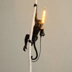 Black monkey on a thick white rope holding a long filament bulb that glows, in front of a plain beige wall; from Clérachic.