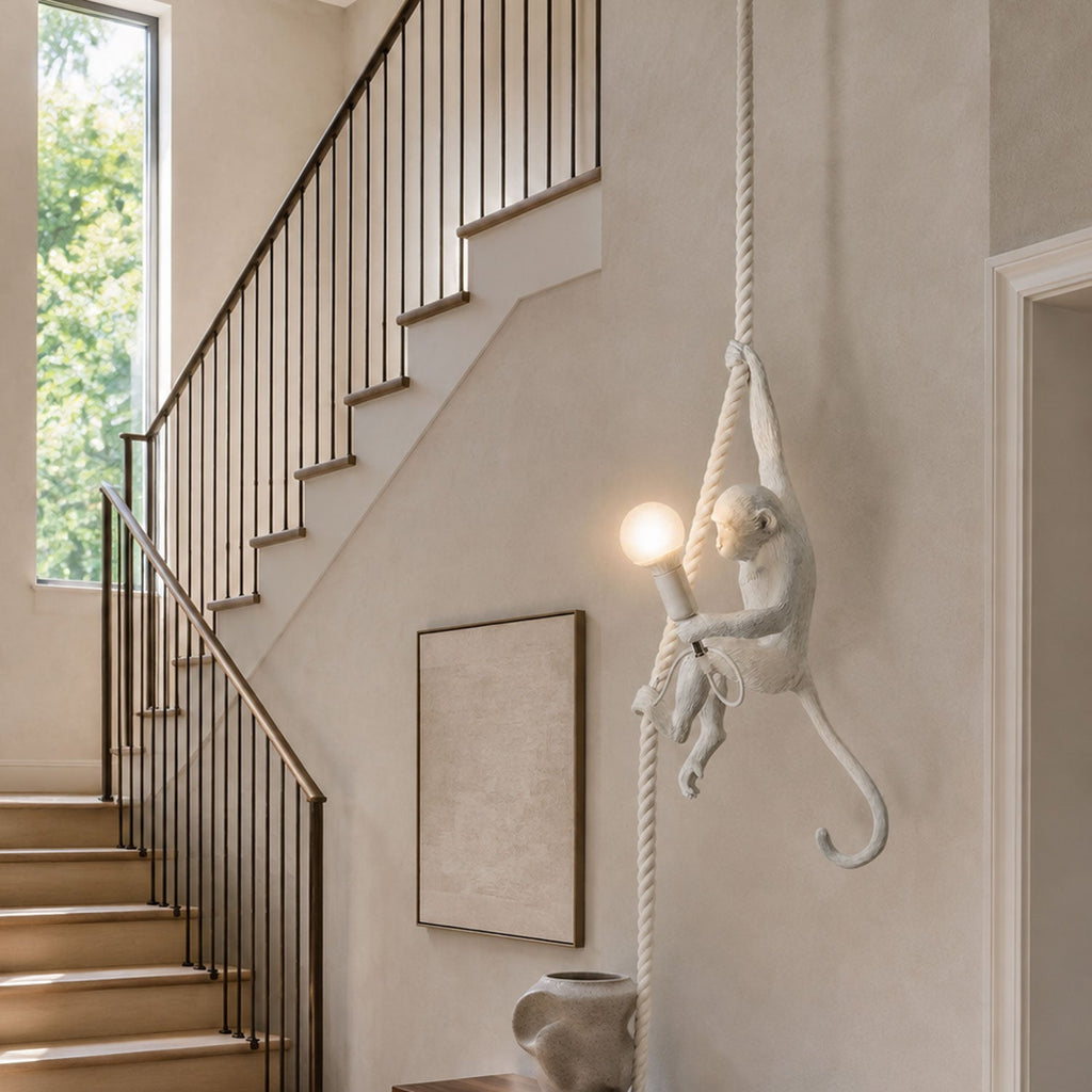 White monkey pendant holding a lit bulb on a pale rope beside a staircase with a dark rail and a framed neutral print; this Clérachic design.