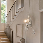 White monkey pendant holding a lit bulb on a pale rope beside a staircase with a dark rail and a framed neutral print; this Clérachic design.