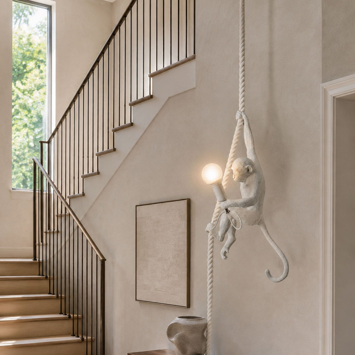 White monkey pendant holding a lit bulb on a pale rope beside a staircase with a dark rail and a framed neutral print; this Clérachic design.