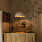 A Clérachic pendant with a ragged gold-lit rim hangs above a weathered cabinet and a rough plaster wall with a fish painting.