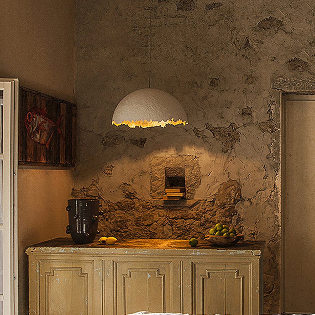 A Clérachic pendant with a ragged gold-lit rim hangs above a weathered cabinet and a rough plaster wall with a fish painting.