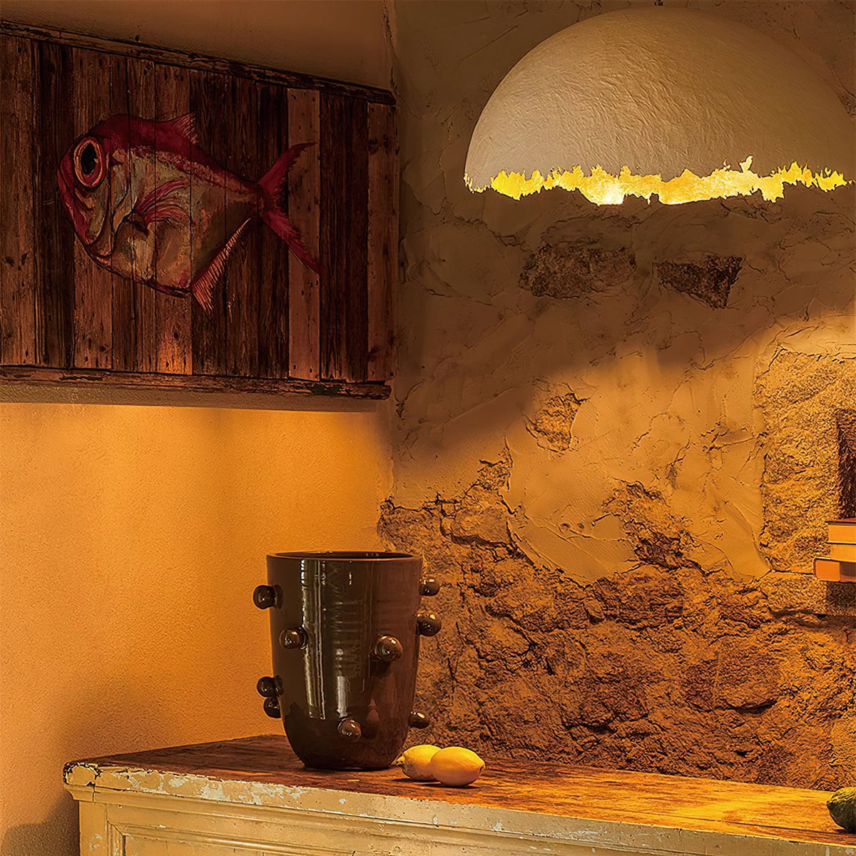 Close view of a Clérachic dome pendant's ragged gold rim against a rough plaster wall, with a fish painting and a dark vase.