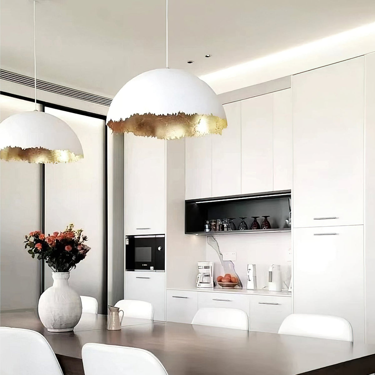 Two Clérachic dome pendants with gold edges hang over a dark dining table in a white kitchen with a black shelf niche.