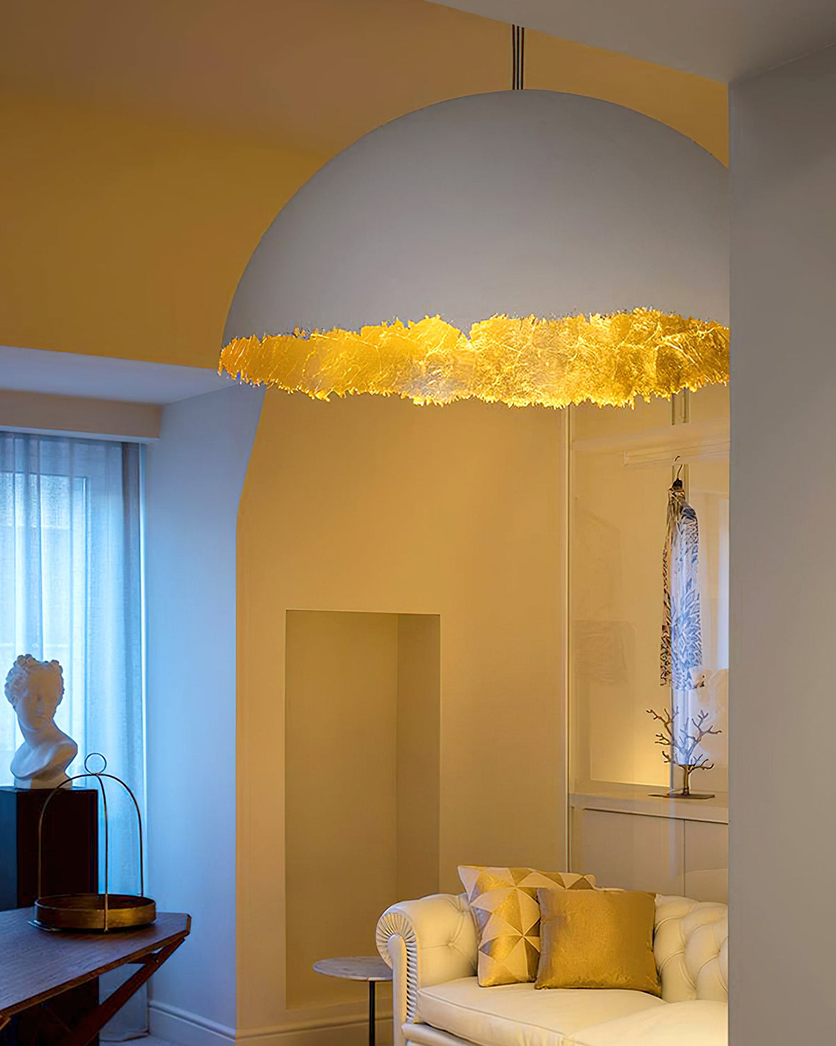 Close view of the white dome and gold-leaf rim of a Clérachic pendant, with a cream sofa and yellow cushions below.