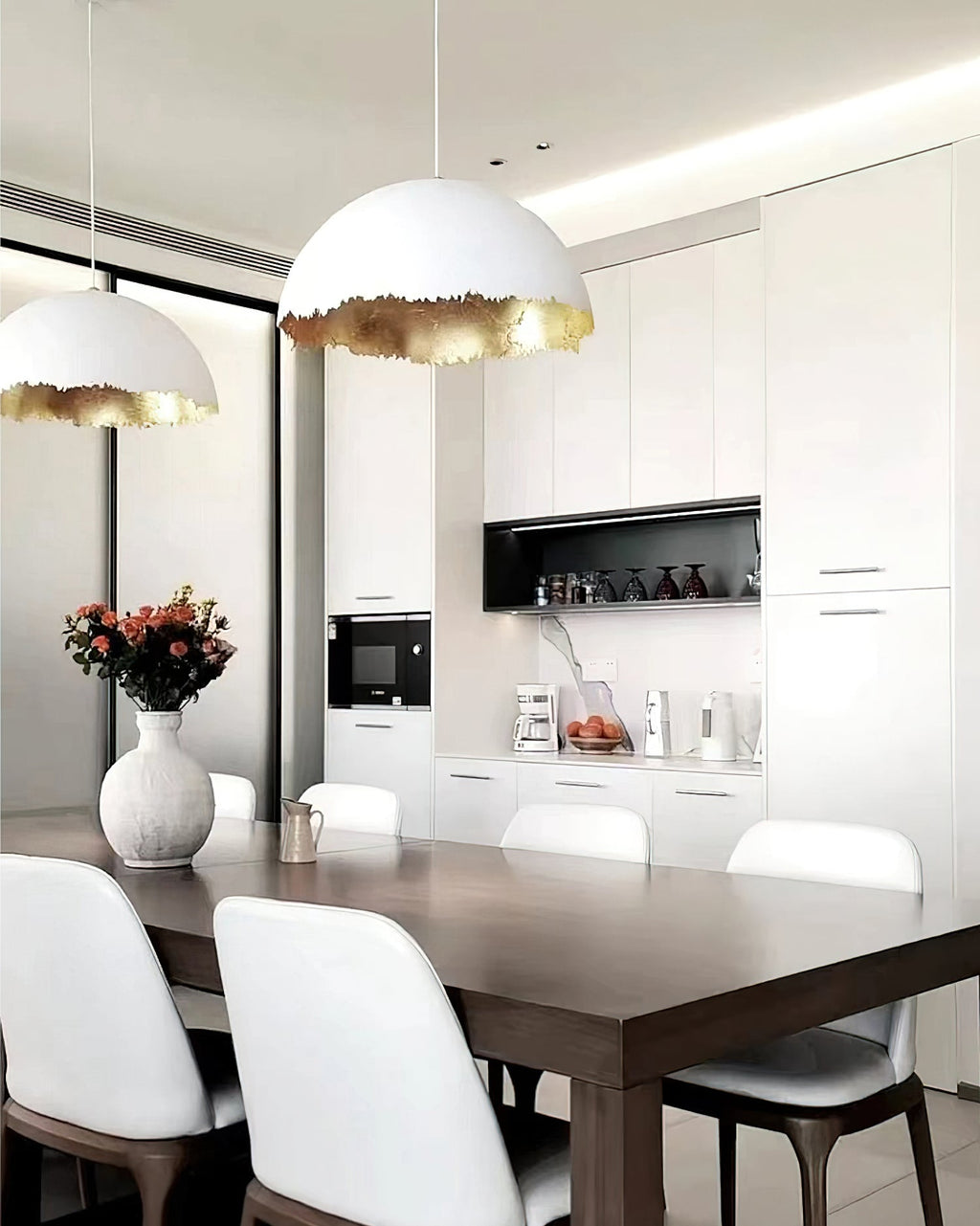 A Clérachic dome pendant hangs over a dining table with white chairs, in front of white cabinets and a vase of flowers.