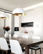 A Clérachic dome pendant hangs over a dining table with white chairs, in front of white cabinets and a vase of flowers.
