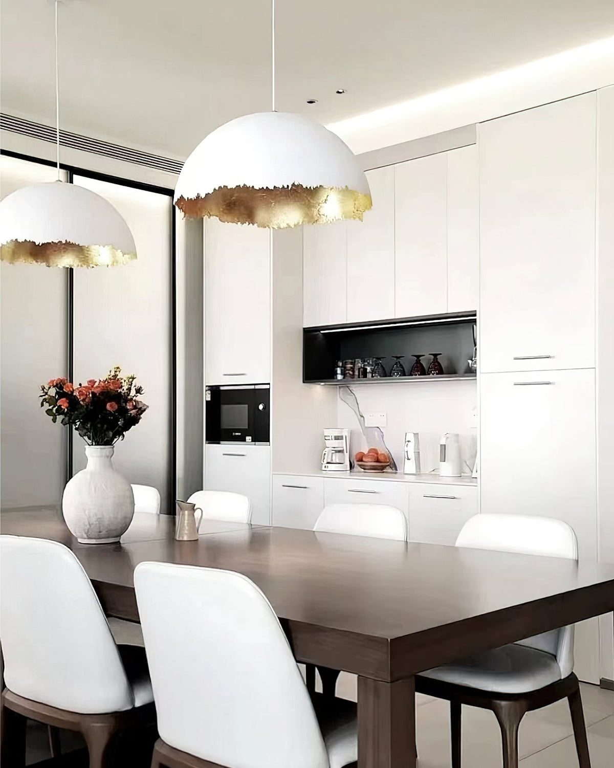 A Clérachic dome pendant hangs over a dining table with white chairs, in front of white cabinets and a vase of flowers.