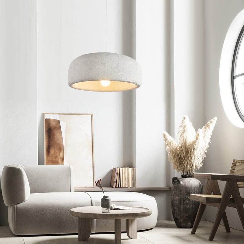 A Clérachic dome pendant hangs in a pale living room above a curved grey sofa, a round coffee table and a vase of dried pampas grass.