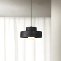 Black stepped-drum Clérachic pendant hanging in front of floor-length white curtains, its round bulb glowing below the shade.
