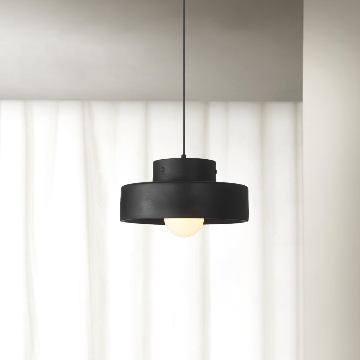 Black stepped-drum Clérachic pendant hanging in front of floor-length white curtains, its round bulb glowing below the shade.