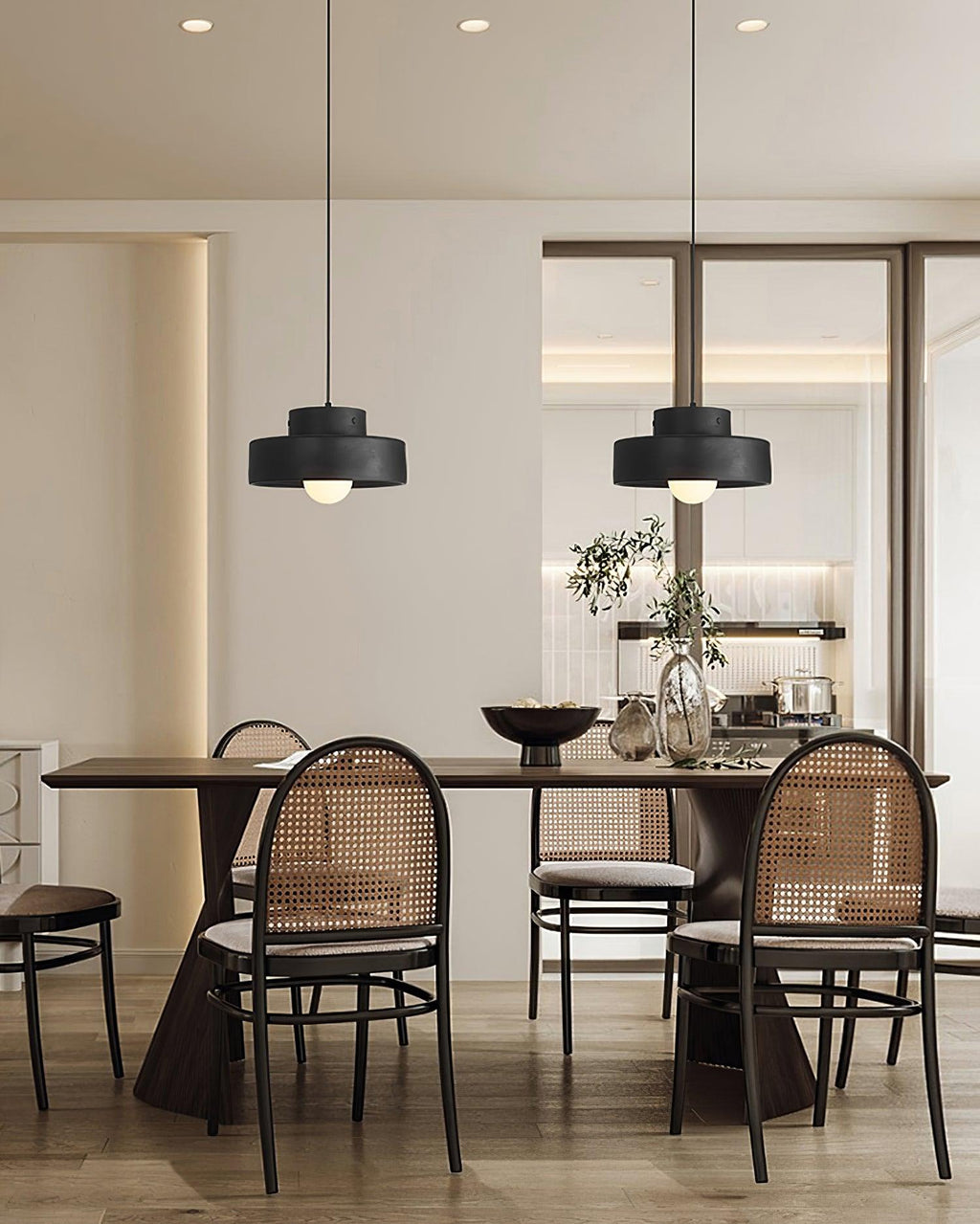 Two black Clérachic pendants in a row over a long dark dining table, with round-backed cane chairs and a plant in a bright, open room.