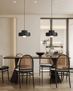 Two black Clérachic pendants in a row over a long dark dining table, with round-backed cane chairs and a plant in a bright, open room.