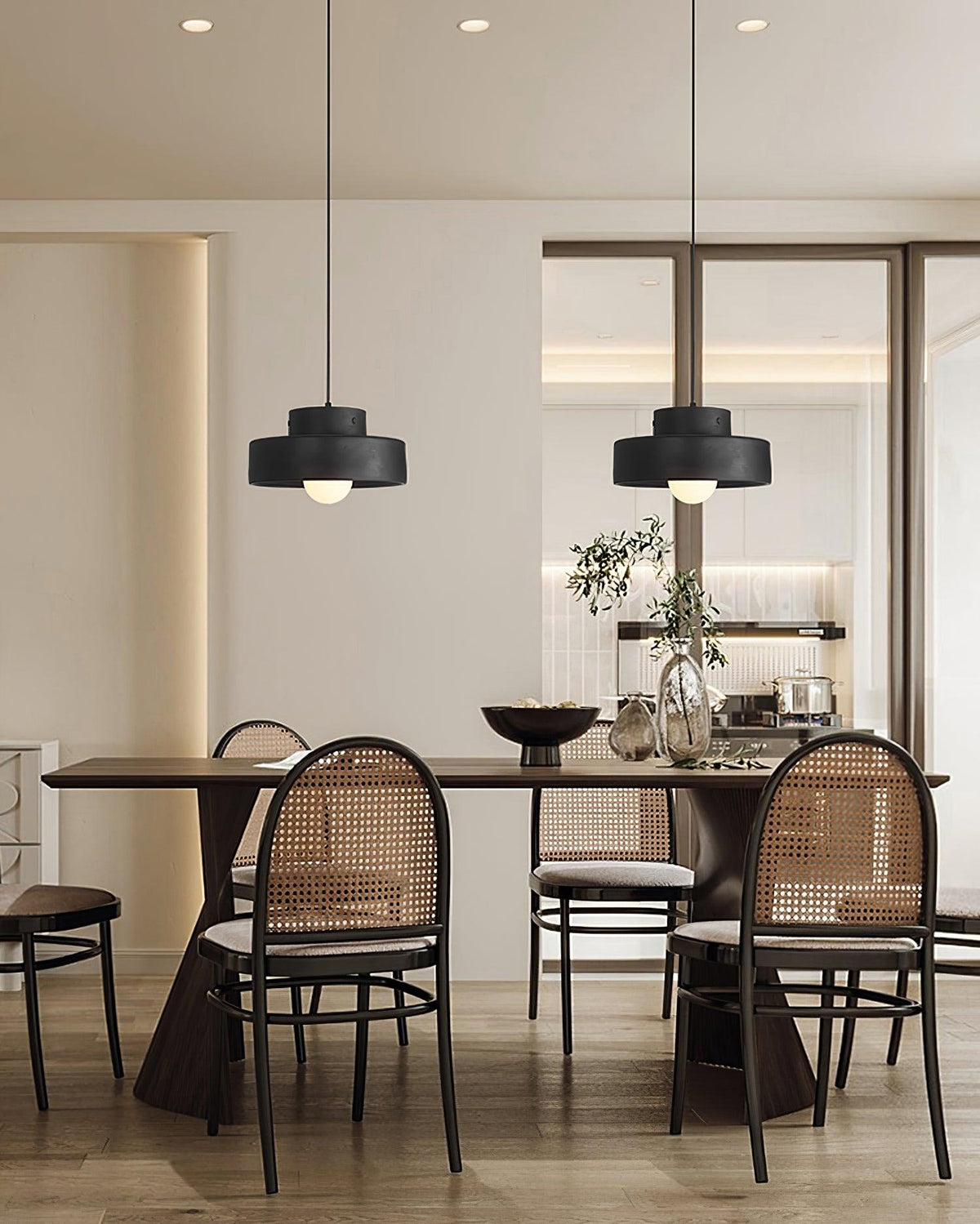 Two black Clérachic pendants in a row over a long dark dining table, with round-backed cane chairs and a plant in a bright, open room.