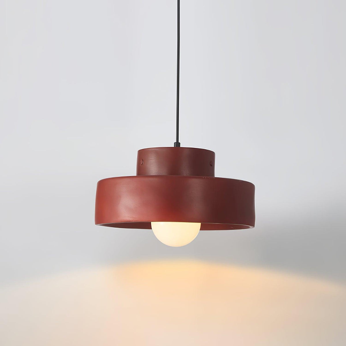A red Clérachic pendant lit against a pale grey wall, throwing a warm pool of light below its stepped resin shade.