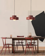 Two red Clérachic pendants over a long wooden table with red chairs, against a pale wall and a large black abstract shape.