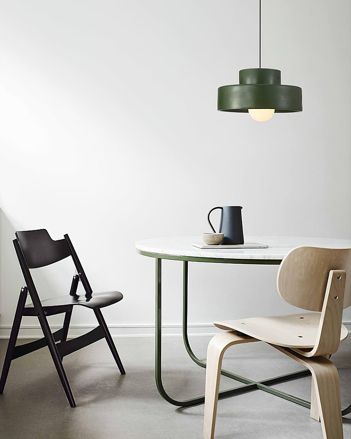 Green Clérachic pendant on a white wall above a round marble-topped table, a folding chair and a moulded plywood chair.