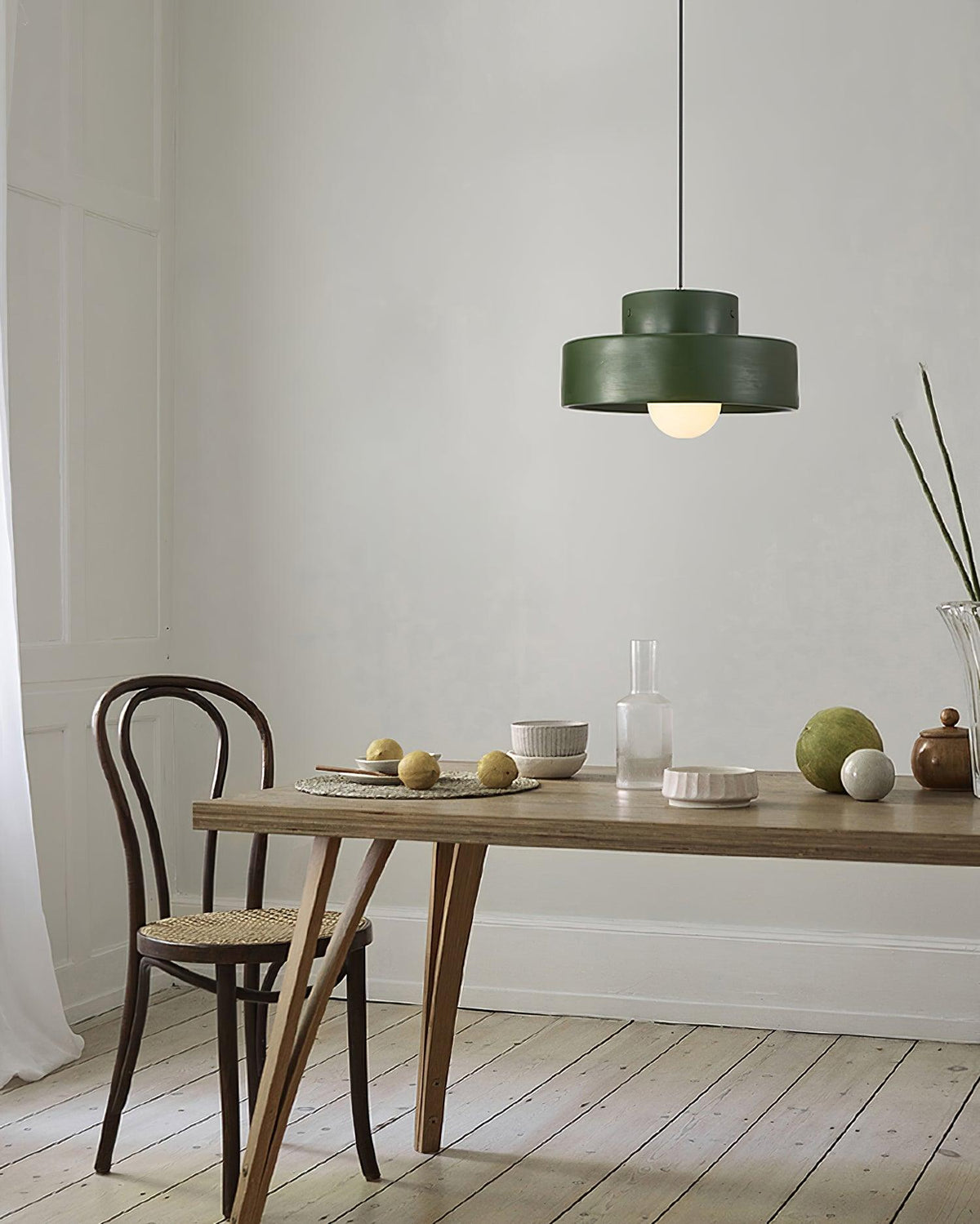 A green Clérachic pendant in a pale room, hung over a wooden table laid with small bowls, a glass bottle and a bentwood chair.