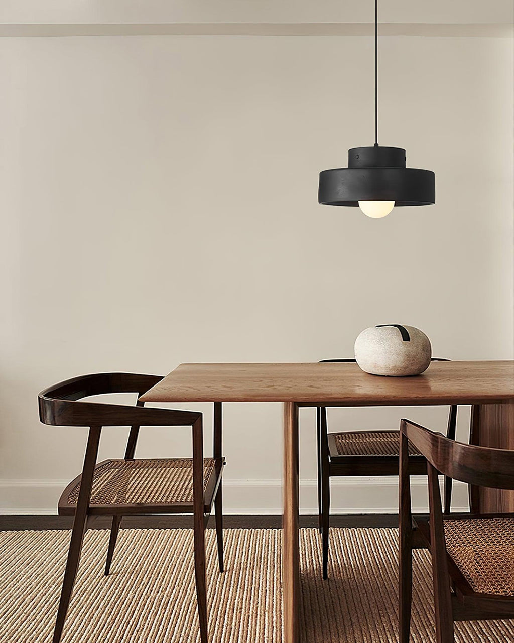 Black Clérachic pendant over a walnut dining table with a pale stone vase, set on a woven rug beside dark cane-backed chairs.