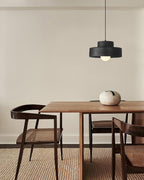 Black Clérachic pendant over a walnut dining table with a pale stone vase, set on a woven rug beside dark cane-backed chairs.