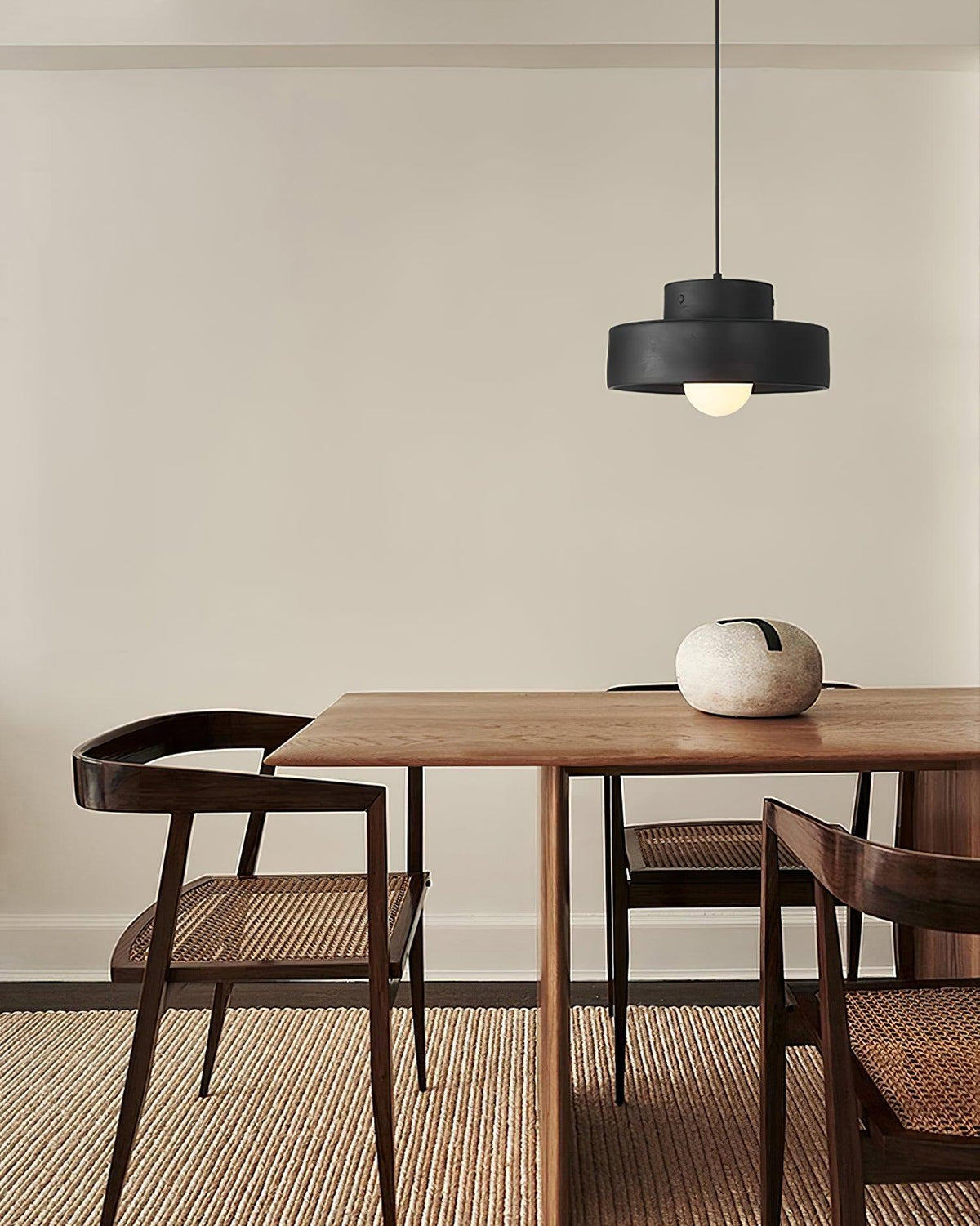 Black Clérachic pendant over a walnut dining table with a pale stone vase, set on a woven rug beside dark cane-backed chairs.