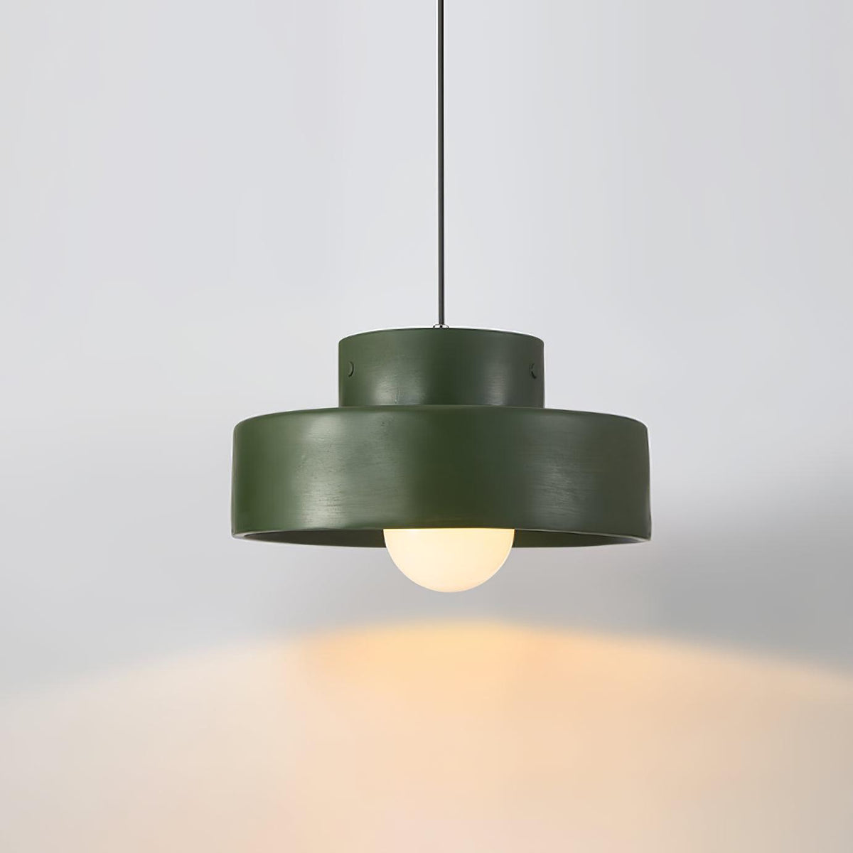 A green Clérachic pendant lit against a soft grey wall, with a pool of warm light on the surface below.