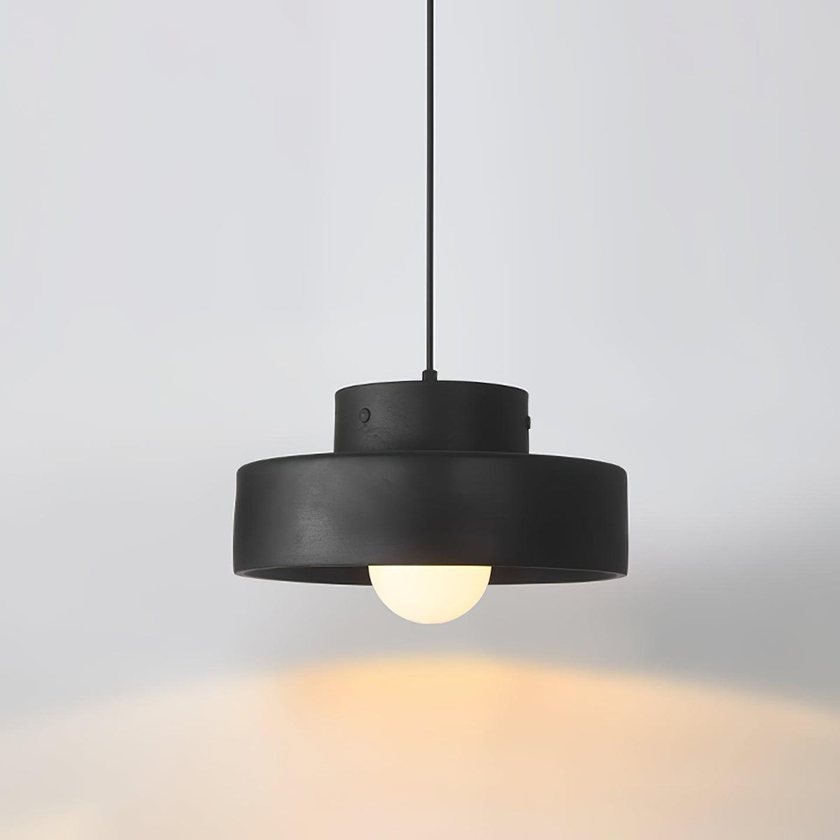 Black Clérachic pendant lit against a soft grey wall, its stepped shade and round bulb casting warm light below.