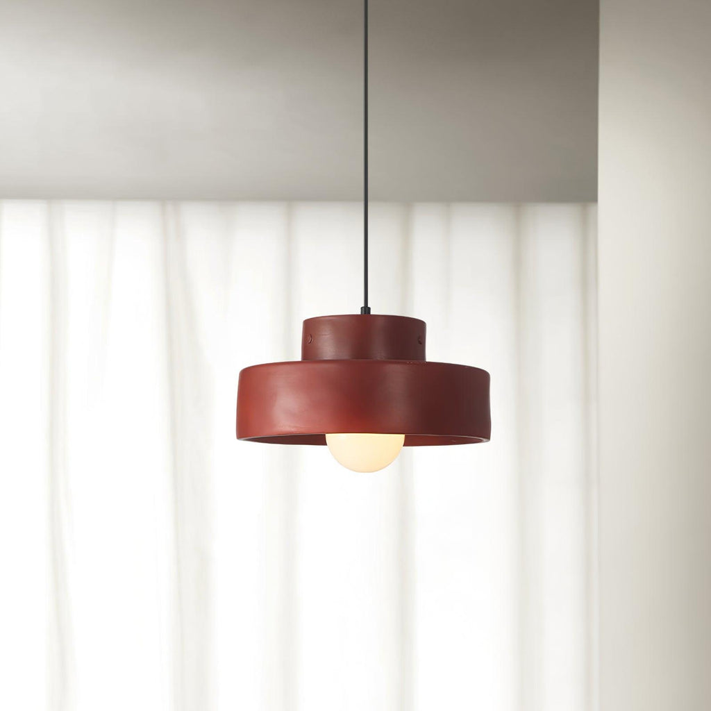 A red Clérachic pendant with its bulb lit, hanging in front of sheer white curtains beside a pale wall.