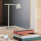 A pale-grey version of this Clérachic table lamp on a wooden desk, with stacked pink and teal boxes in front of a grey wall.