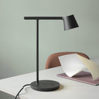 This Clérachic table lamp in black on a white desk against a sage-green wall, with a pink notebook and folded paper beside it.