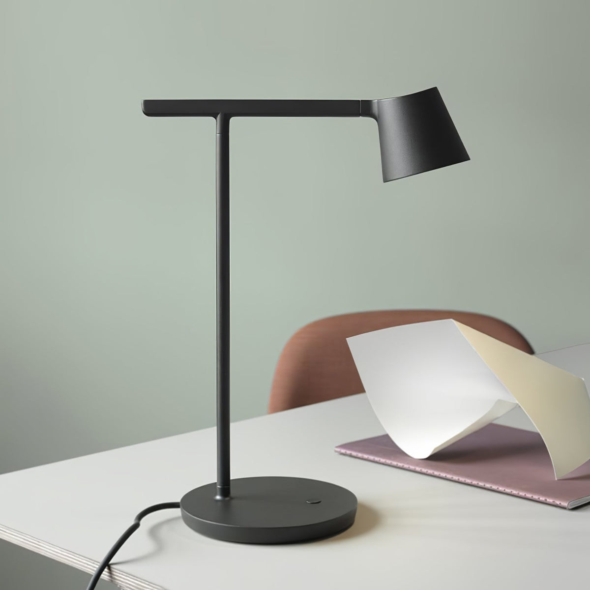 This Clérachic table lamp in black on a white desk against a sage-green wall, with a pink notebook and folded paper beside it.