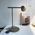 An olive-green Clérachic table lamp lit on a white desk, its head tilted up beside a magazine and a framed print.