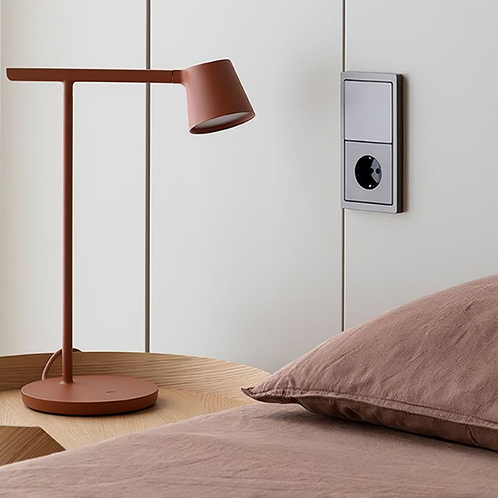 Rust-red Clérachic table lamp on a round wooden bedside table, set against pink bedding and a wall socket.