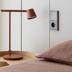 Rust-red Clérachic table lamp on a round wooden bedside table, set against pink bedding and a wall socket.