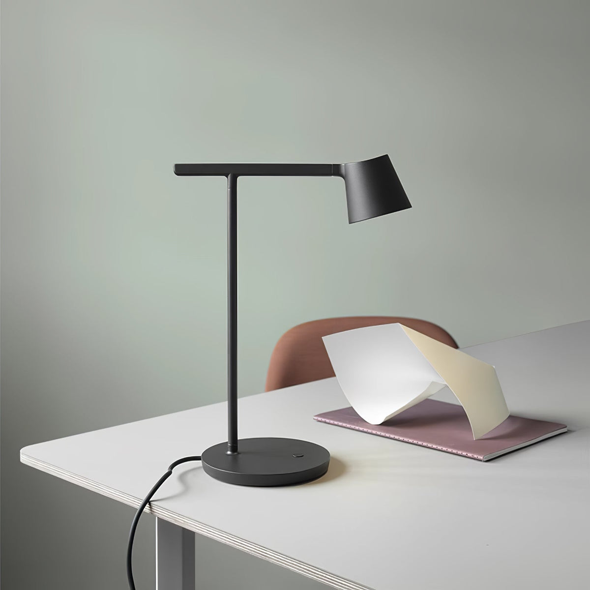 Black Clérachic table lamp on a white desk in front of a soft green wall, its cord trailing down the edge of the desk.