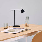 A black Clérachic table lamp on a light oak table beside two glass carafes and an open book.