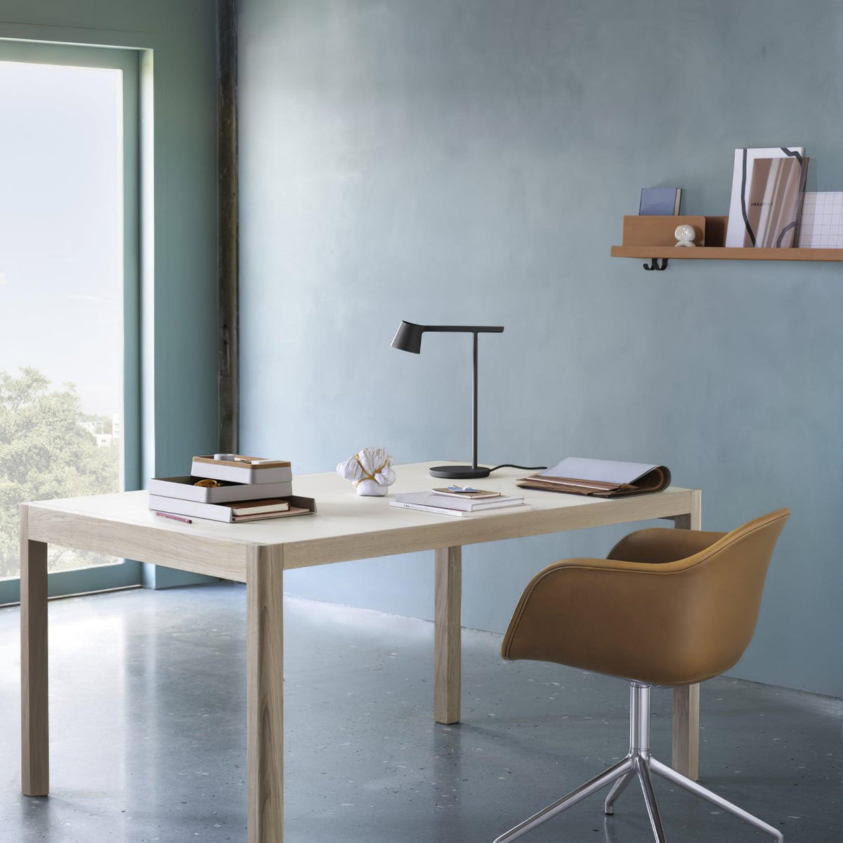 Small black Clérachic table lamp on a desk across a blue-grey room, with a shelf of books above and an open window beside it.