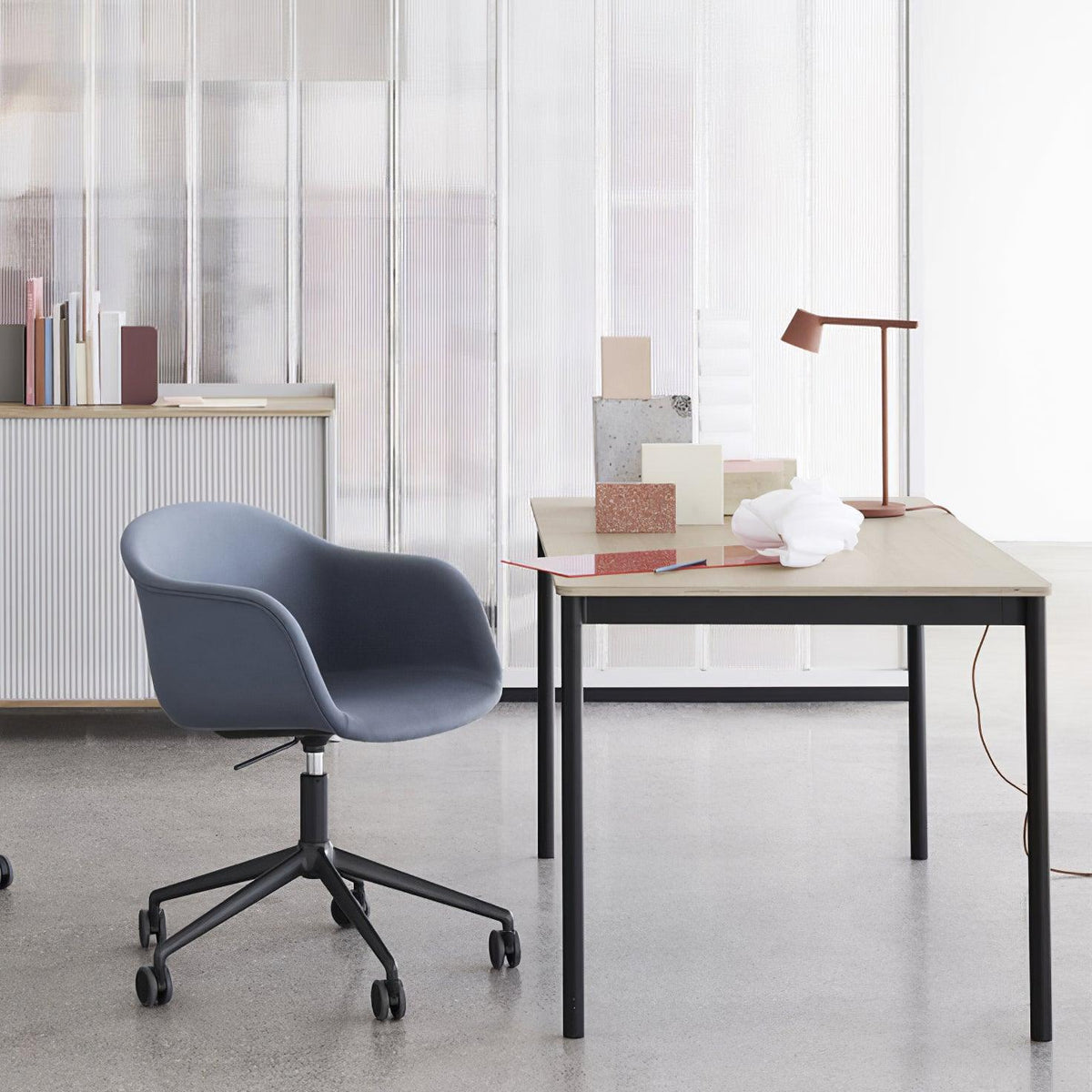 Distant view of a rust-red Clérachic table lamp on a desk in a bright studio, with a blue swivel chair in front.