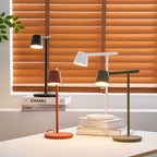 This Clérachic table lamp in four finishes, with three heads lit, in front of orange blinds on a white desk.