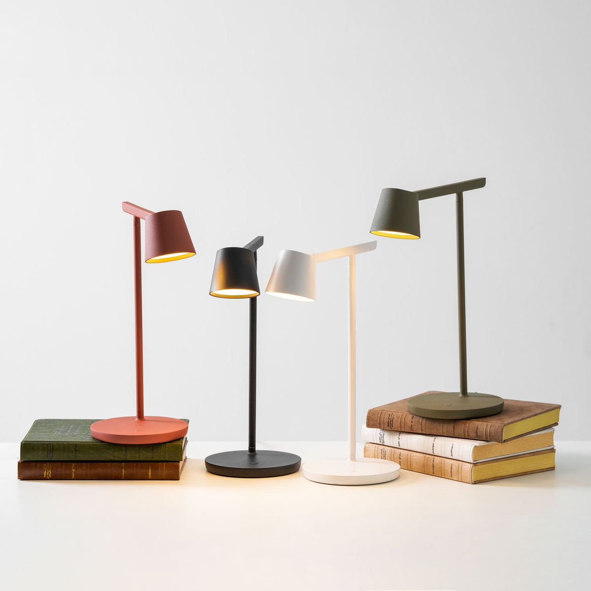 Four Clérachic table lamps on a white surface, two of them resting on stacks of old books, all with lit heads.