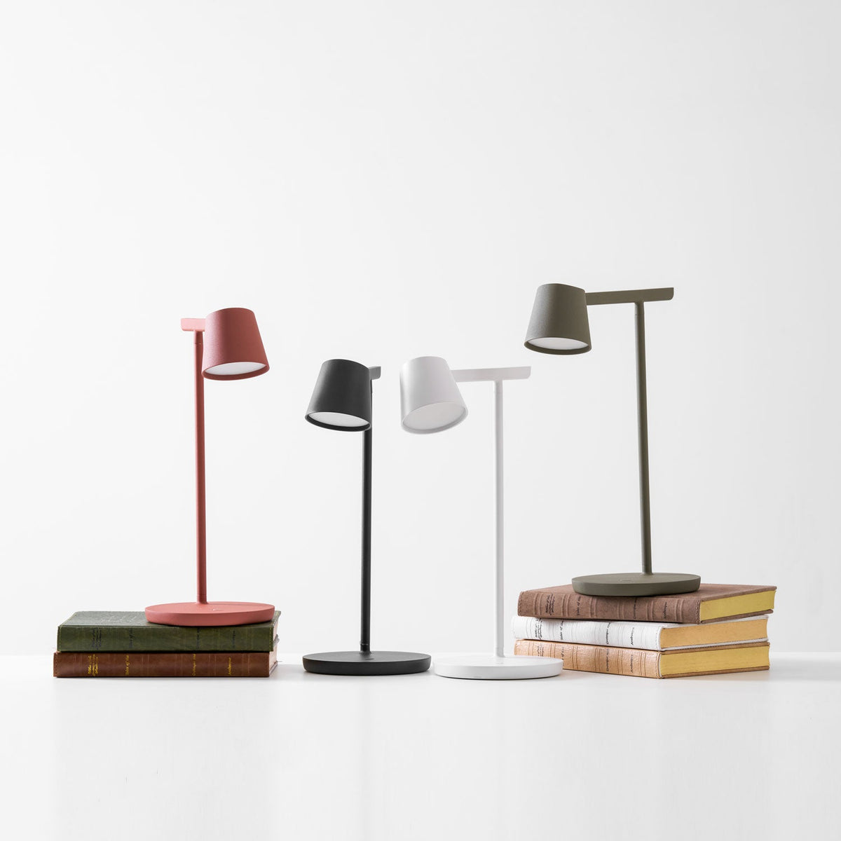 A group of four Clérachic table lamps on white, with the red and olive lamps on stacks of cloth-bound books.