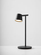 A black Clérachic table lamp lit, with a gold-toned inside to the shade and a round base on a pale grey backdrop.