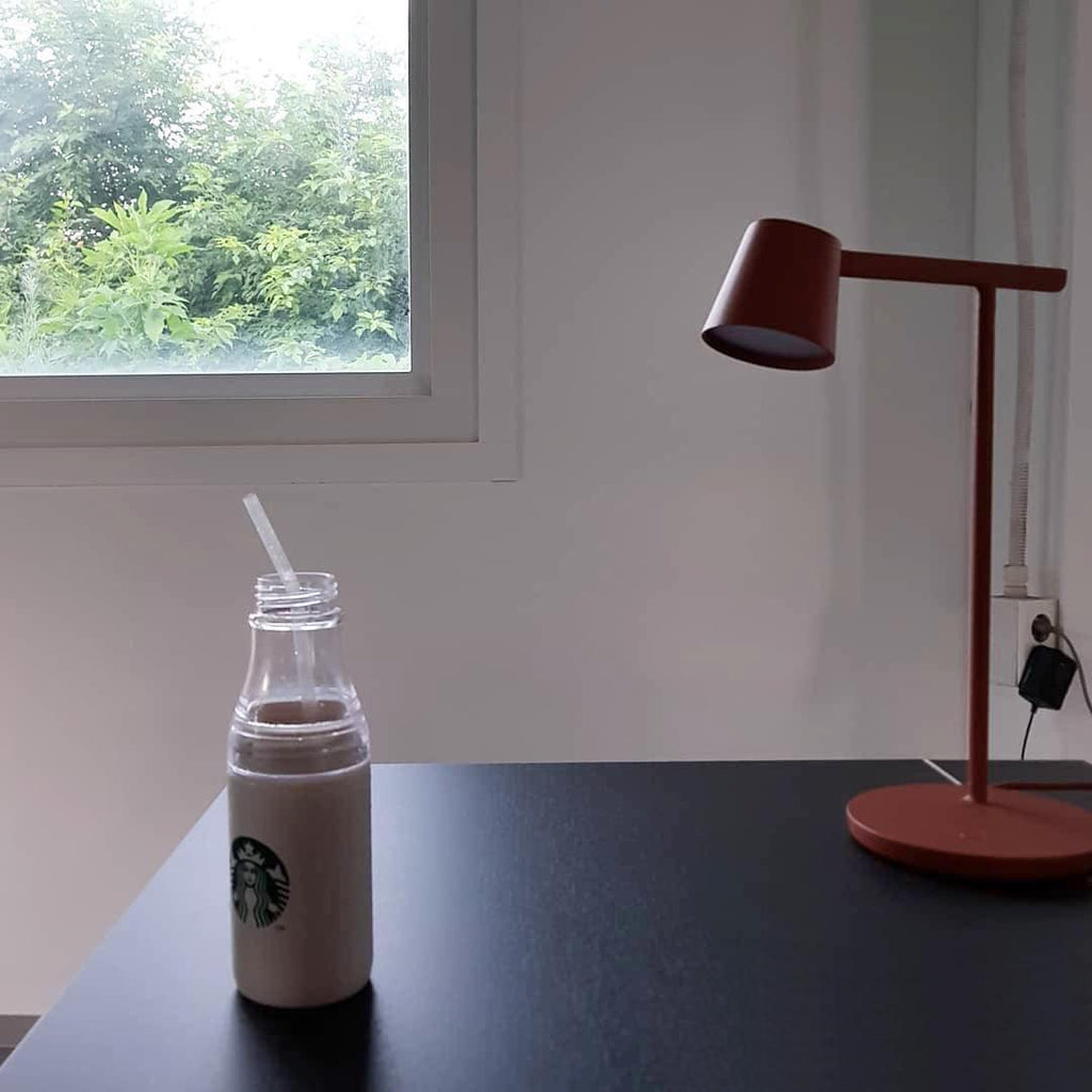 Rust-red Clérachic table lamp on a black desk beside a sunny window, with a bottle with a straw in the foreground.