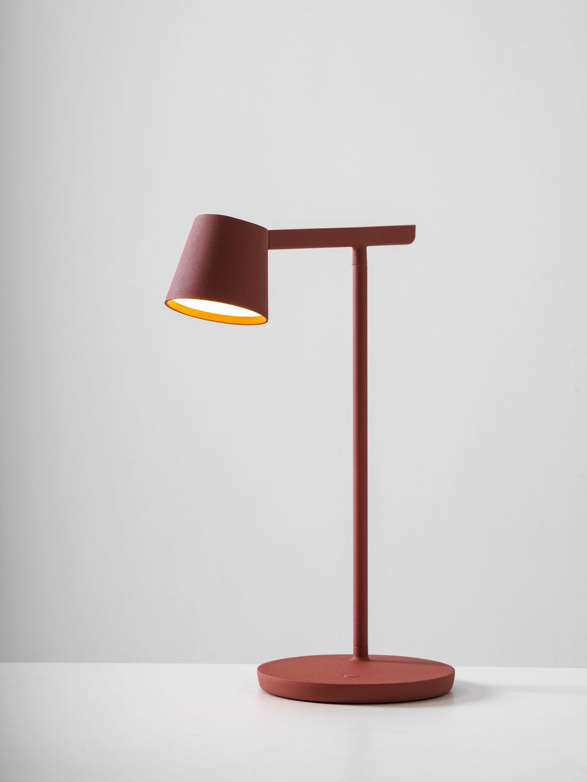 A red Clérachic table lamp with its head lit and a warm gold inner shade, on a pale grey backdrop.