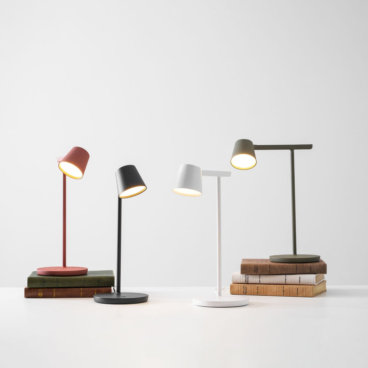 Four Clérachic table lamps in red, black, white and olive, with the red and olive ones on stacks of books and all heads lit.