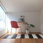 A small dark Clérachic table lamp on a white desk in a bright room, with a red chair, a striped rug and a potted monstera.