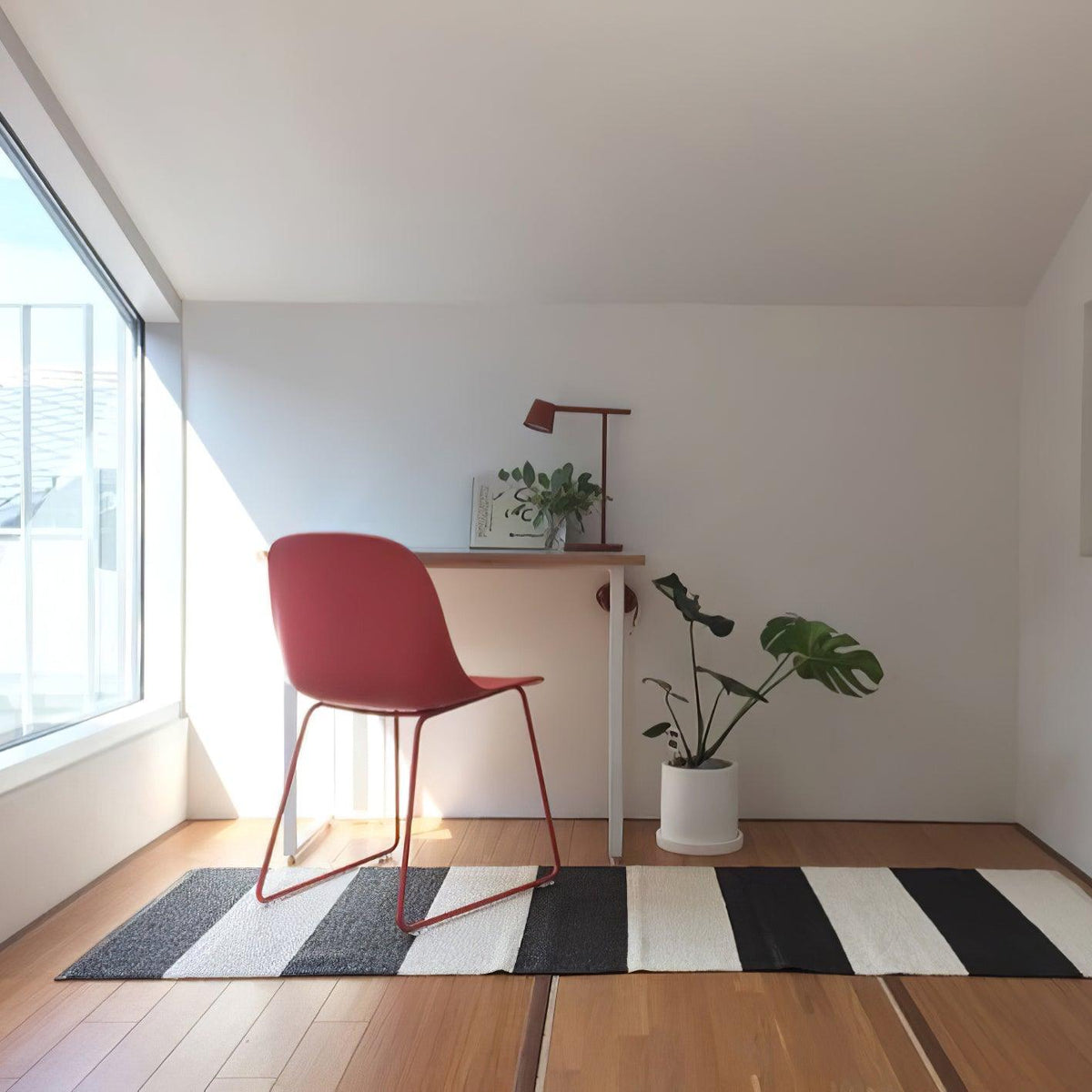 A small dark Clérachic table lamp on a white desk in a bright room, with a red chair, a striped rug and a potted monstera.