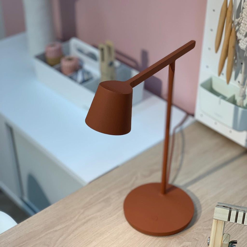 Close view of the angled head of a rust-red Clérachic table lamp above a wooden desk with a blurred organiser behind.